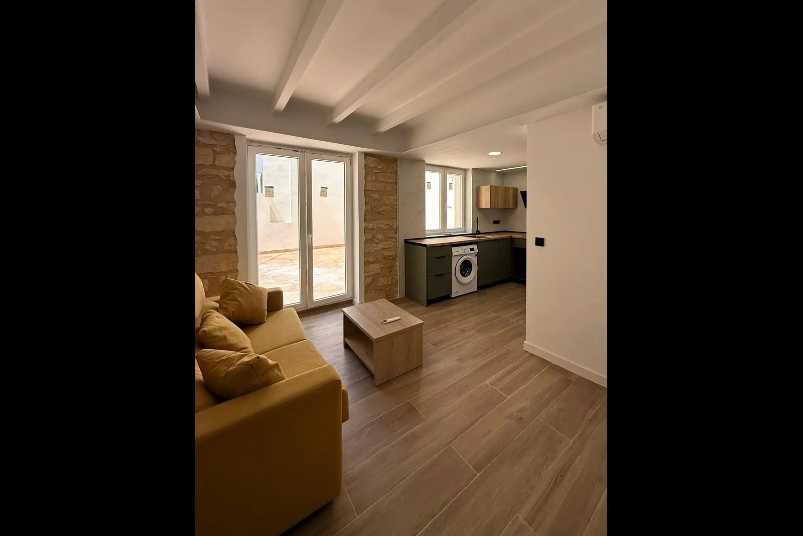 Salón-cocina reformado con pilares de piedra vista, techo de vigas blancas y salida a la terraza; cocina verde con encimera de madera