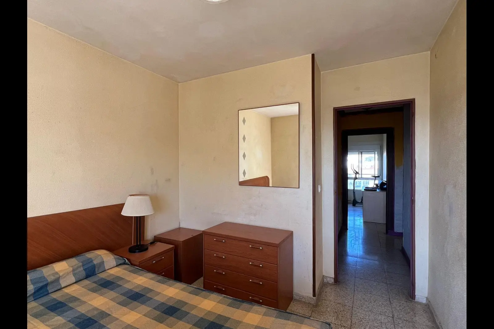 Dormitorio principal sin reformar, con cabecero y cómoda de madera, espejo cuadrado y suelo de terrazo; al fondo, el pasillo