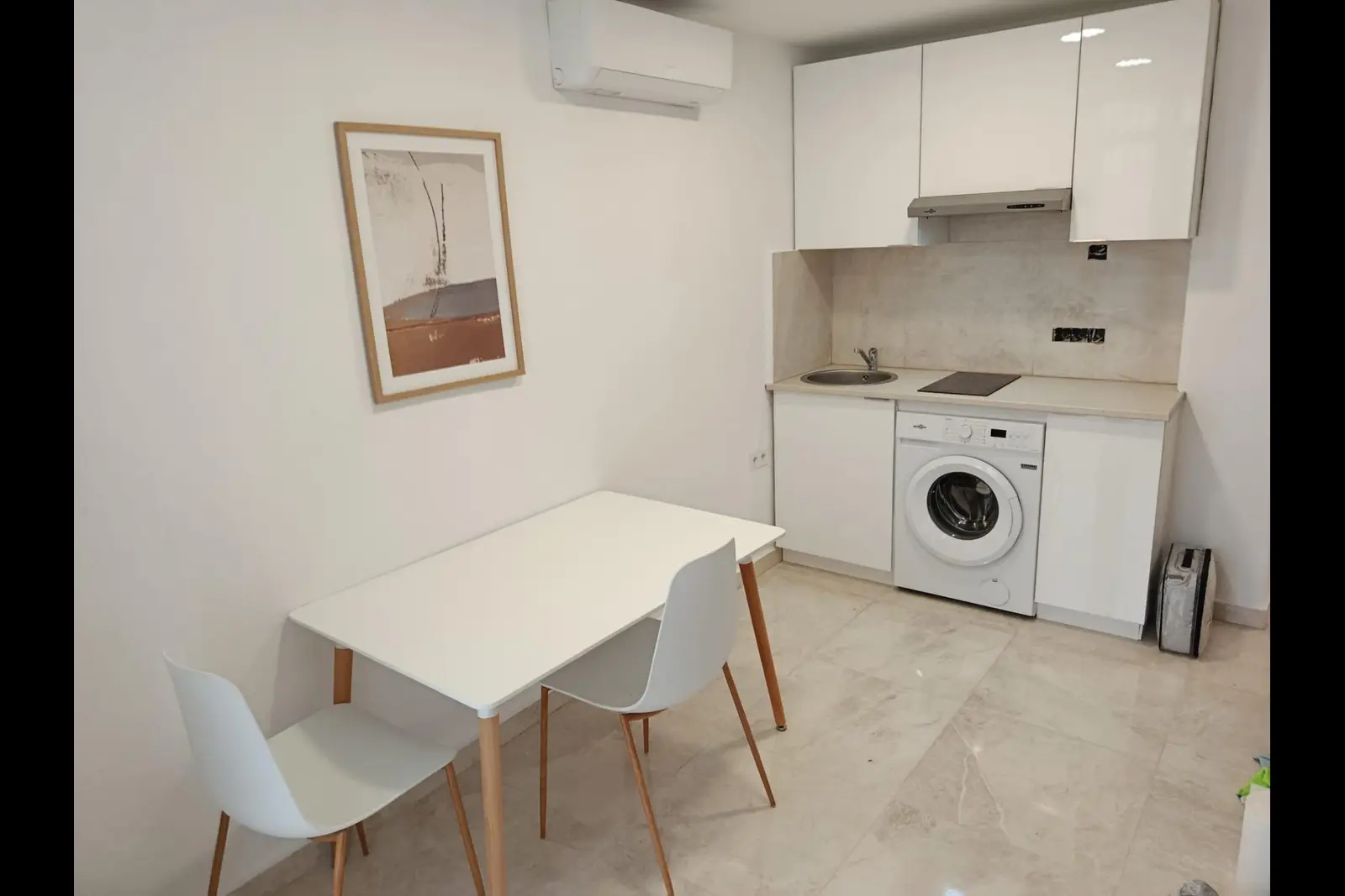 Salón-cocina reformado, con cocina blanca, lavadora integrada, mesa para dos y suelo porcelánico claro