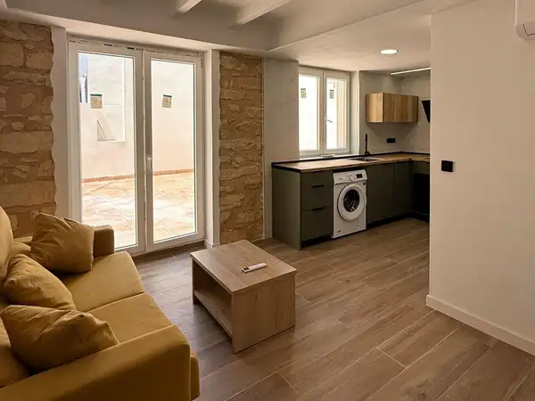 Salón-cocina reformado con pilares de piedra vista, techo de vigas blancas y salida a la terraza; cocina verde con encimera de madera