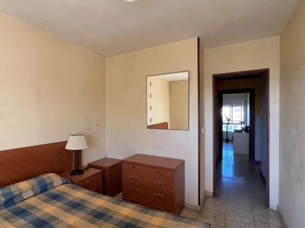 Dormitorio principal sin reformar, con cabecero y cómoda de madera, espejo cuadrado y suelo de terrazo; al fondo, el pasillo